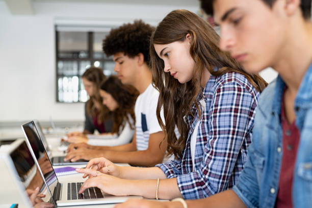 A row of diverse high school or college students, including young men and women, intently working on their laptops in a bright classroom