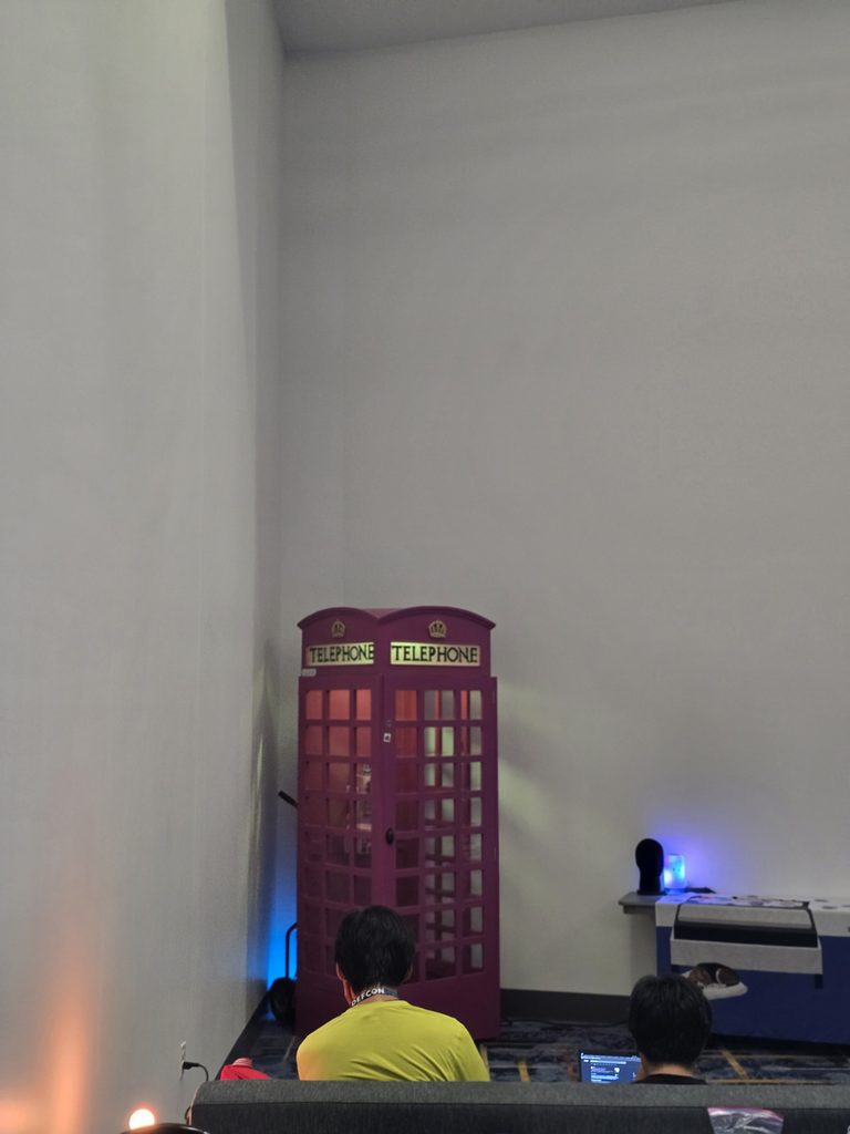 A magenta British-style telephone booth standing in the corner of a large indoor room with high white walls, with people sitting on a couch in the foreground.